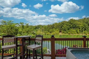 Une terrasse en bois, dotée de chaises et d’un jacuzzi, offre une vue sur un lac, avec une forêt en arrière-plan.