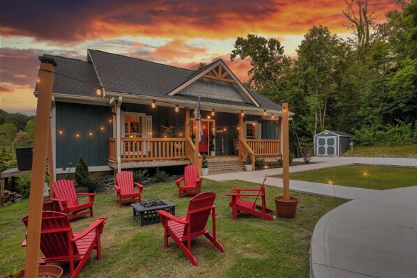 Une maison avec un porche, des chaises Adirondack rouges et un petit abri de jardin dans la cour.