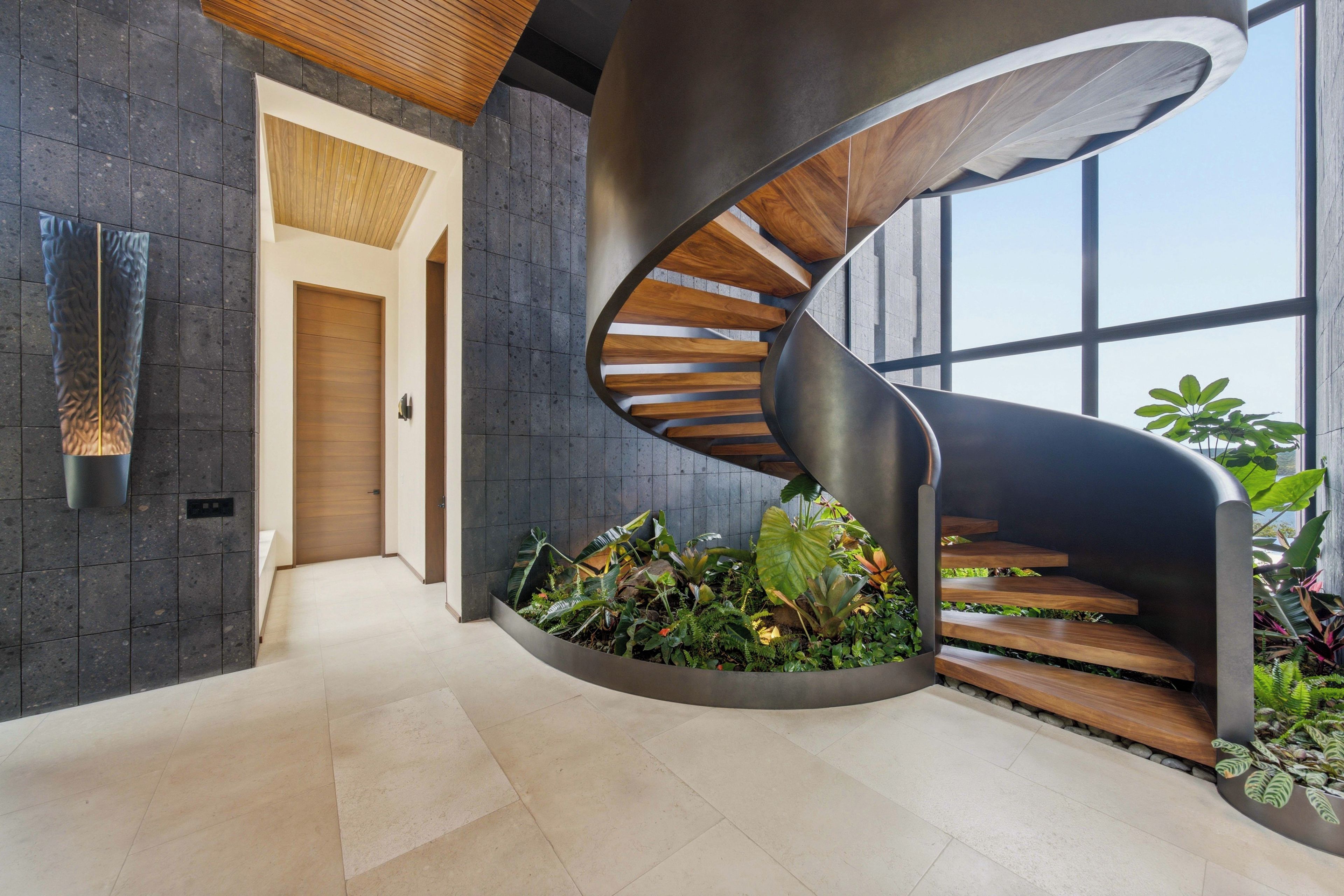 A modern spiral staircase with wooden steps and a metal railing, surrounded by a planter with various plants.