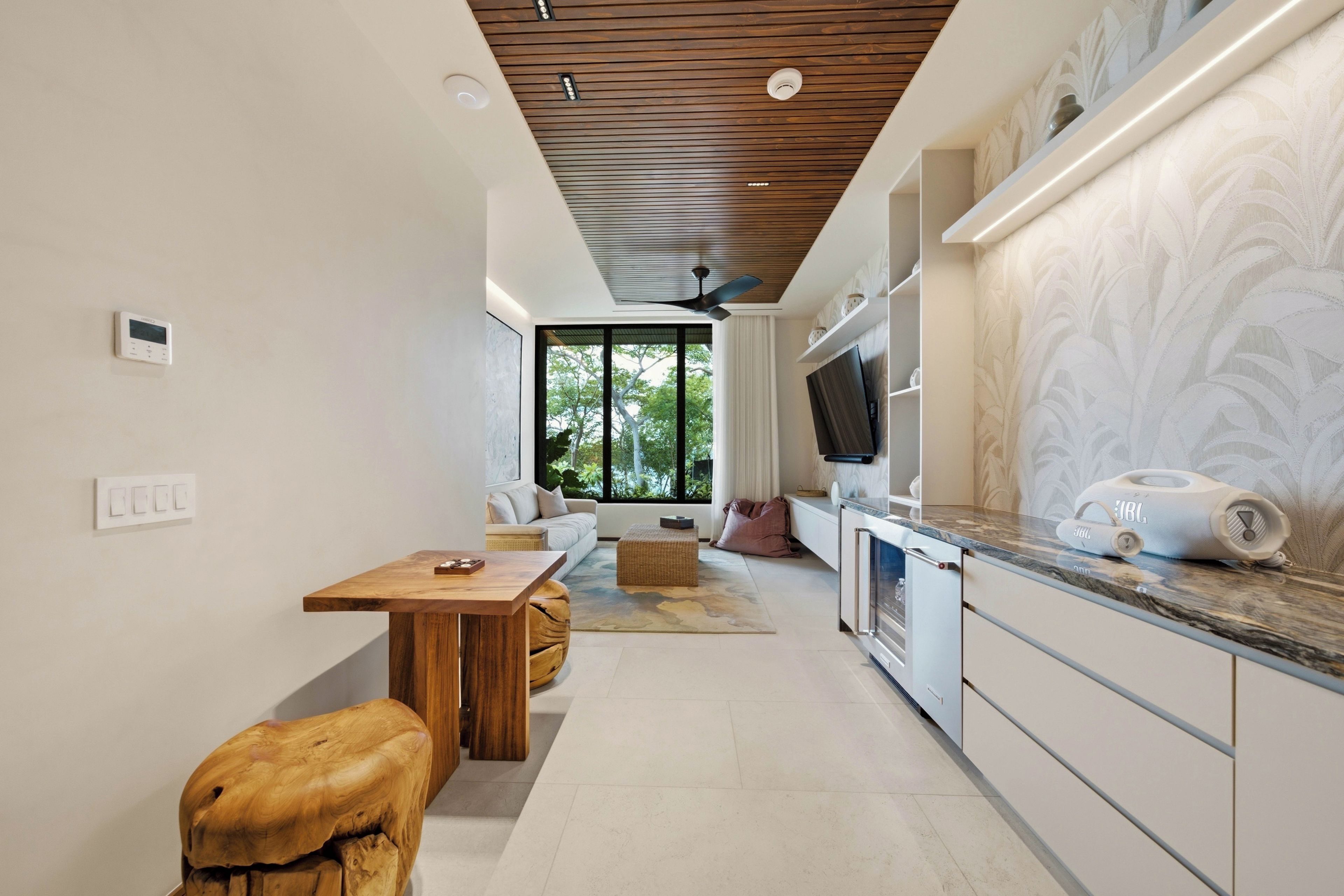 Een moderne keuken met een houten plafond, een eethoek met een tafel en krukken, en een groot raam met uitzicht op groen.