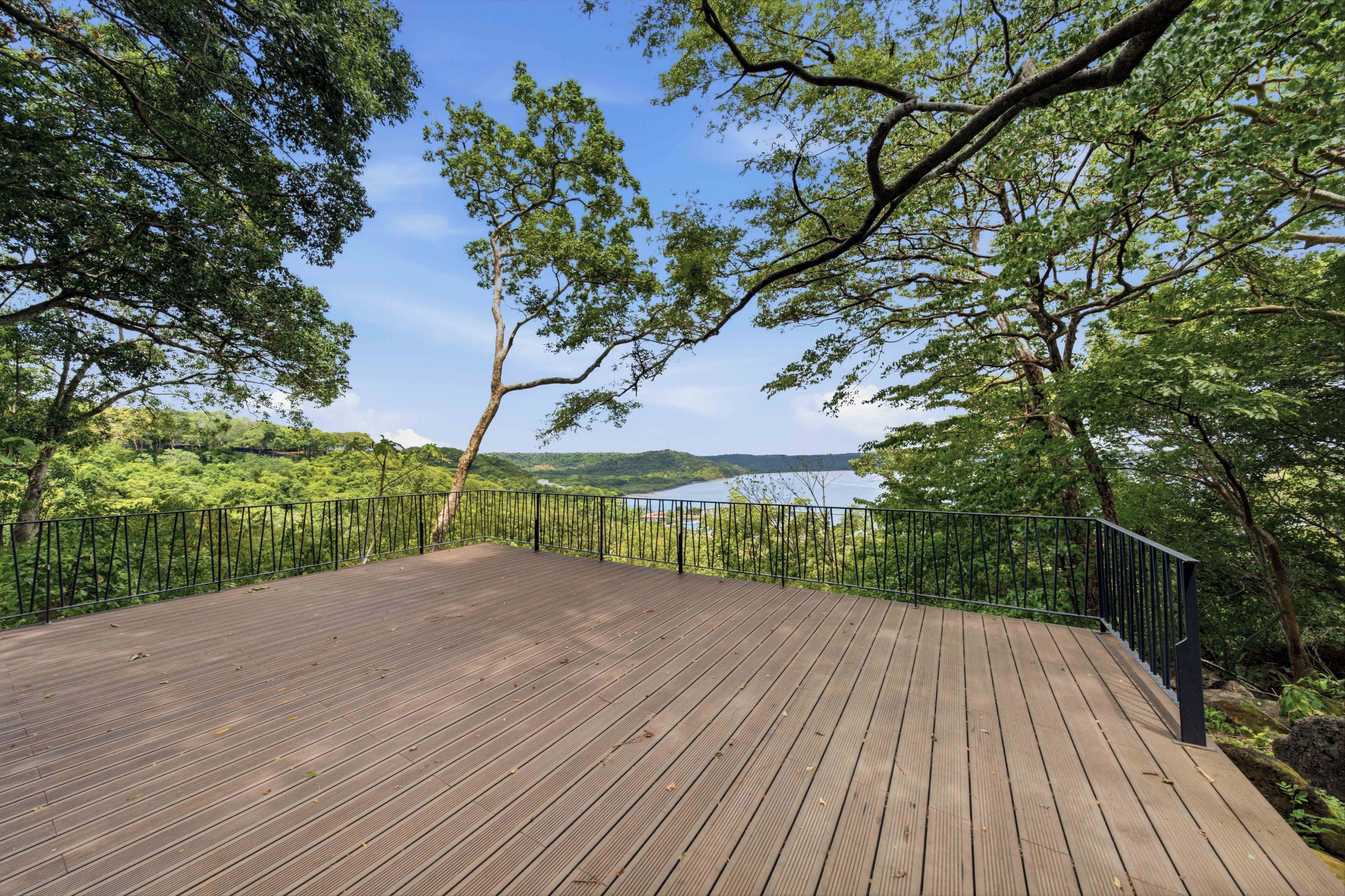 A wooden deck with a metal railing, surrounded by trees and a view of a lake.