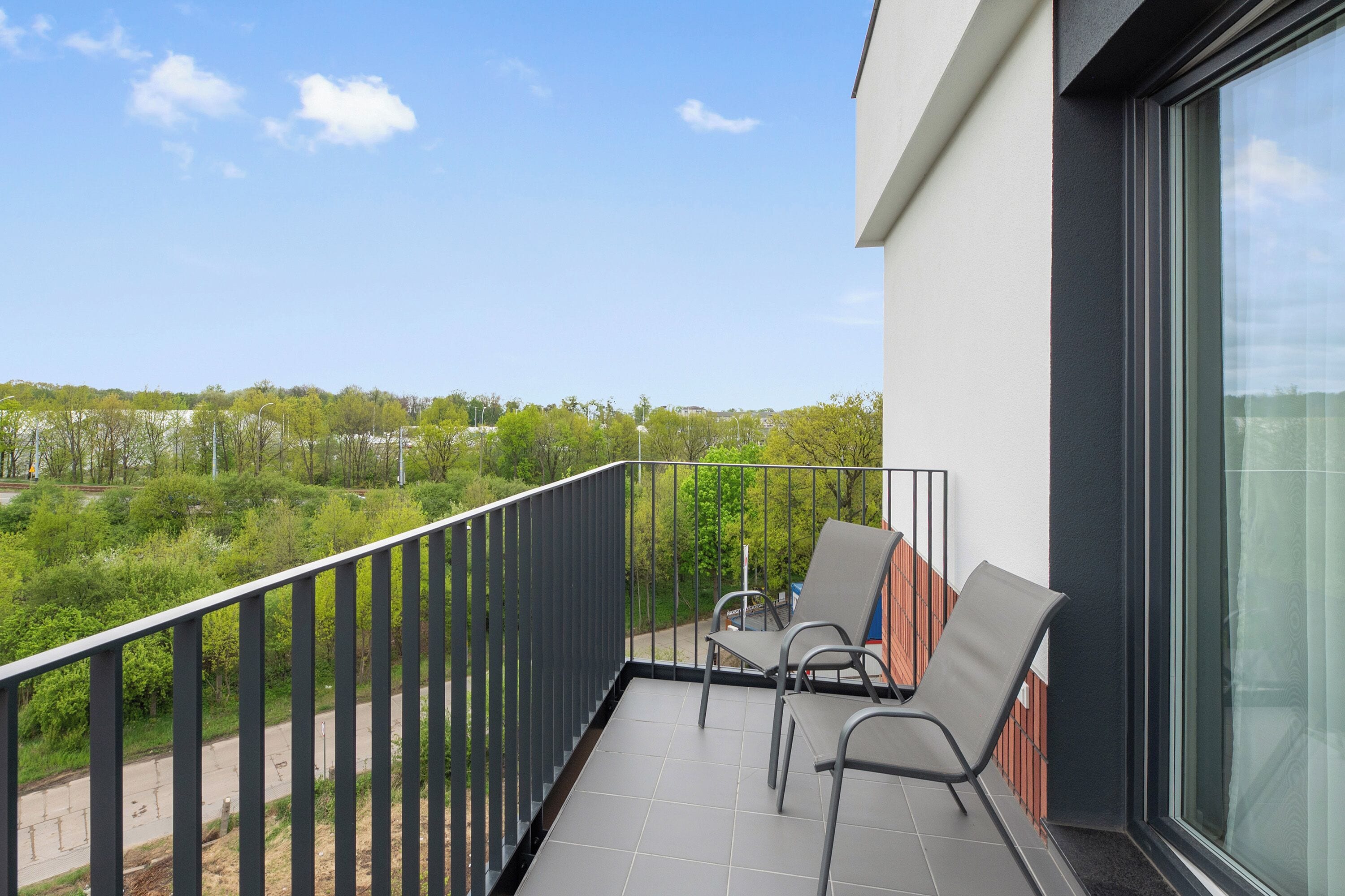 A balcony with two chairs and a table, overlooking a green landscape with trees and buildings.