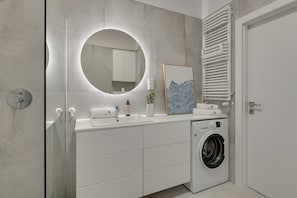 A modern bathroom with a built-in washing machine, a round mirror, and a towel rack. - Faro Beach Apartments with parking (Gdańsk)