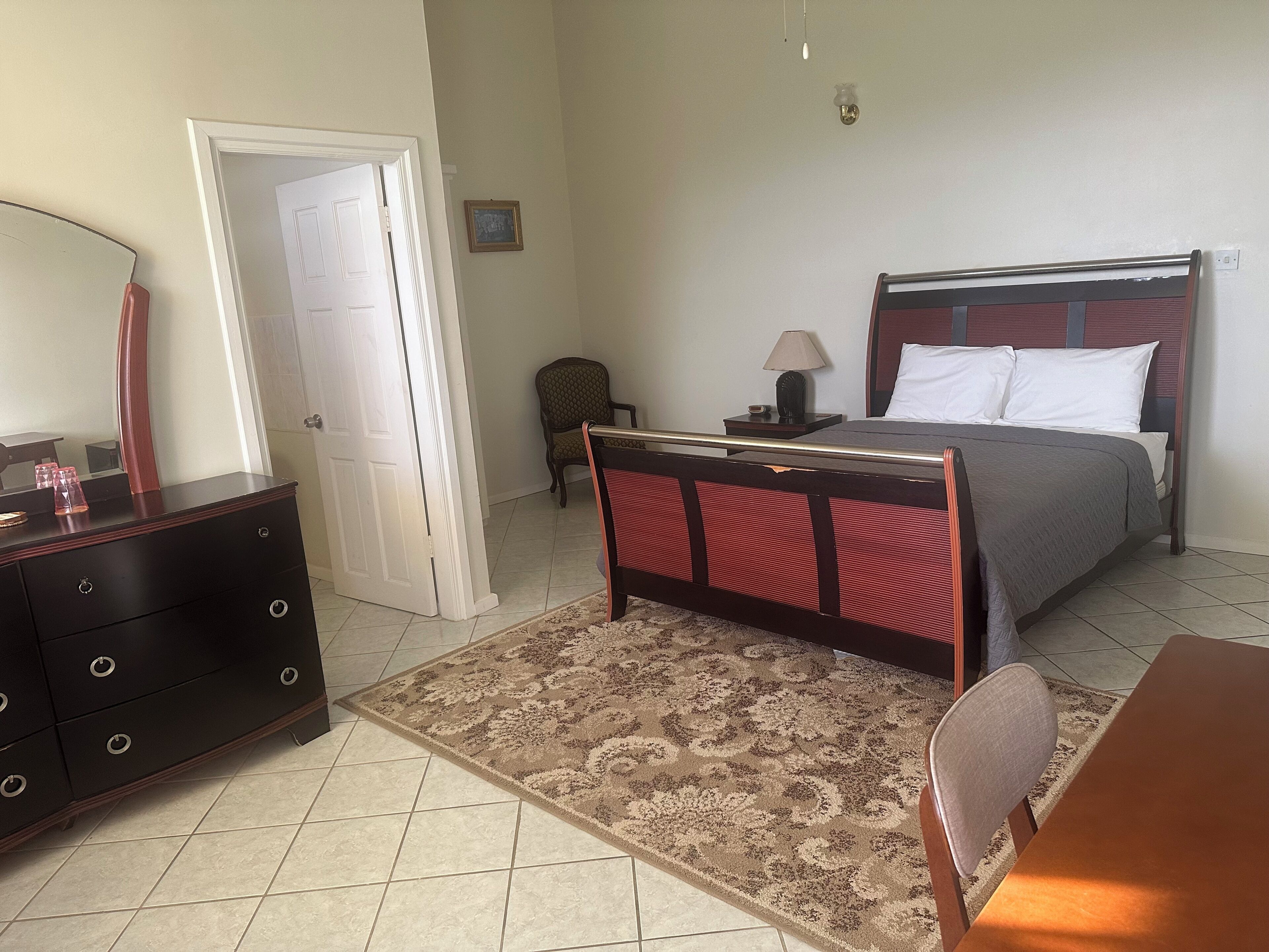 Une chambre à coucher comprenant un lit, une table de chevet, une lampe, une chaise, une commode et une porte.
