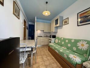 Ein kompakter Wohnbereich mit Küchenzeile, einem Essbereich mit Stühlen, einer mit Blumenmuster bezogenen Couch und einer Deckenleuchte.
