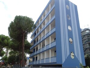 Edifício com várias andares, varandas, placa de estacionamento e árvores ao fundo.