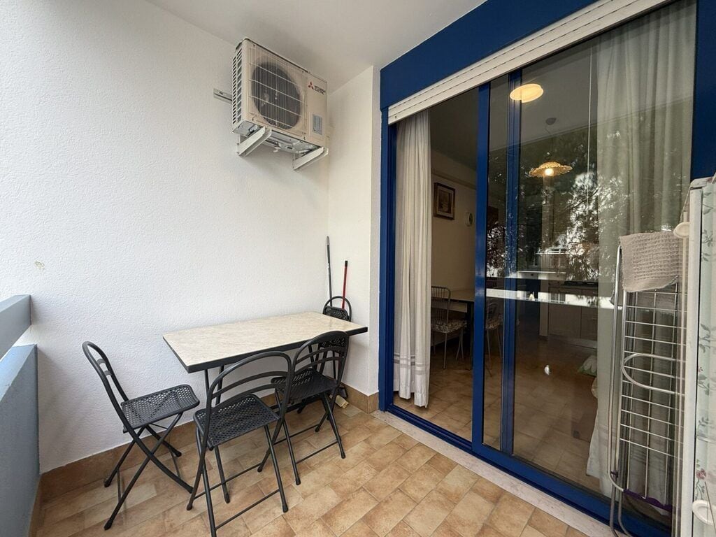 Un balcón con mesa y sillas, un equipo de aire acondicionado y una puerta corredera de cristal que da a un espacio interior.