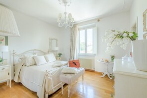Ein Schlafzimmer mit einem Bett, Nachttischen, einer Kommode, einem Fenster mit Vorhängen, einer Heizung und einer Pendelleuchte.