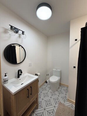 Een badkamer met een wit wastafelmeubel, een zwarte kraan en een ronde spiegel. Er is een toilet met een wit deksel en een zwarte toiletborstel. De vloer heeft een zwart-wit geometrisch patroon.