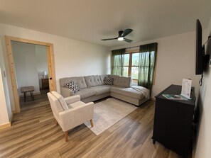 Una sala de estar con un sofá modular, un sillón individual, piso de madera, ventilador de techo, ventana con cortinas y un televisor de pantalla plana sobre un pedestal.