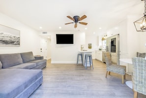 A modern living room with a grey sofa, a ceiling fan, a flat-screen TV, and a kitchen area with a fridge and bar stools. - Great 4 Families, Walk 2 Beach and Bay W/Parking! (Newport Beach)