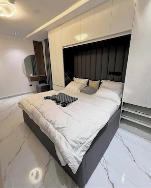 A modern bedroom with a large bed, built-in headboard, and a vanity with a mirror. - Hossana Apartment (Port Harcourt)