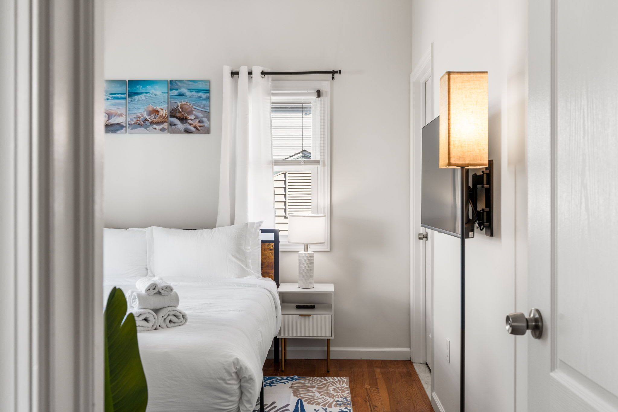 A hotel room with a bed, a bedside table with a lamp, a window with blinds, and a wall-mounted TV.