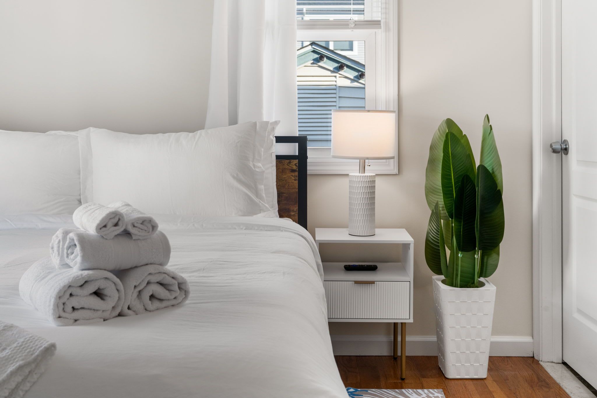 A neatly made bed with rolled towels, a bedside table with a lamp, and a potted plant.