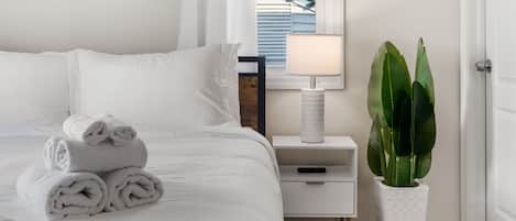 A neatly made bed with rolled towels, a bedside table with a lamp, and a potted plant.