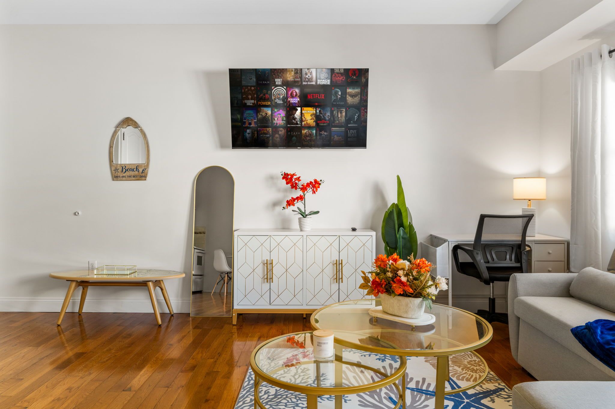 A modern living room with a wooden floor, a glass coffee table, a sofa, a TV mounted on the wall, and a cabinet with decorative items.