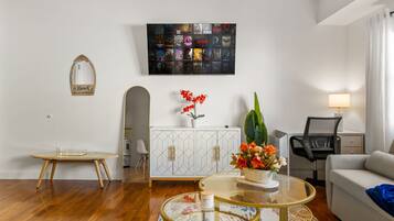 A modern living room with a wooden floor, a glass coffee table, a sofa, a TV mounted on the wall, and a cabinet with decorative items.