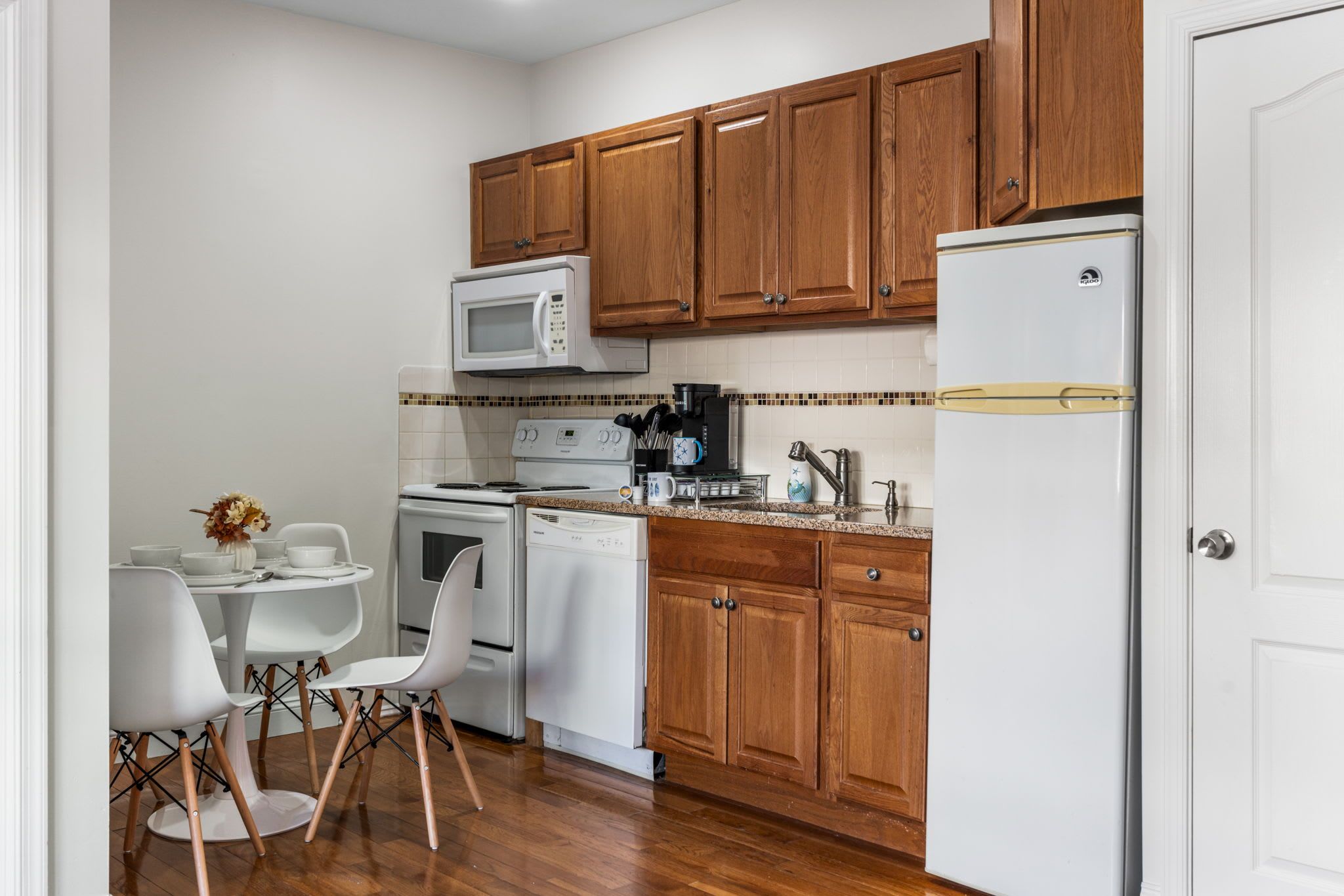 A compact kitchen with wooden cabinets, a white refrigerator, a microwave, a stove, and a dishwasher. There is a dining table with chairs and a vase of dried flowers.