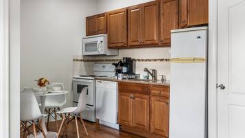 A compact kitchen with wooden cabinets, a white refrigerator, a microwave, a stove, and a dishwasher. There is a dining table with chairs and a vase of dried flowers.