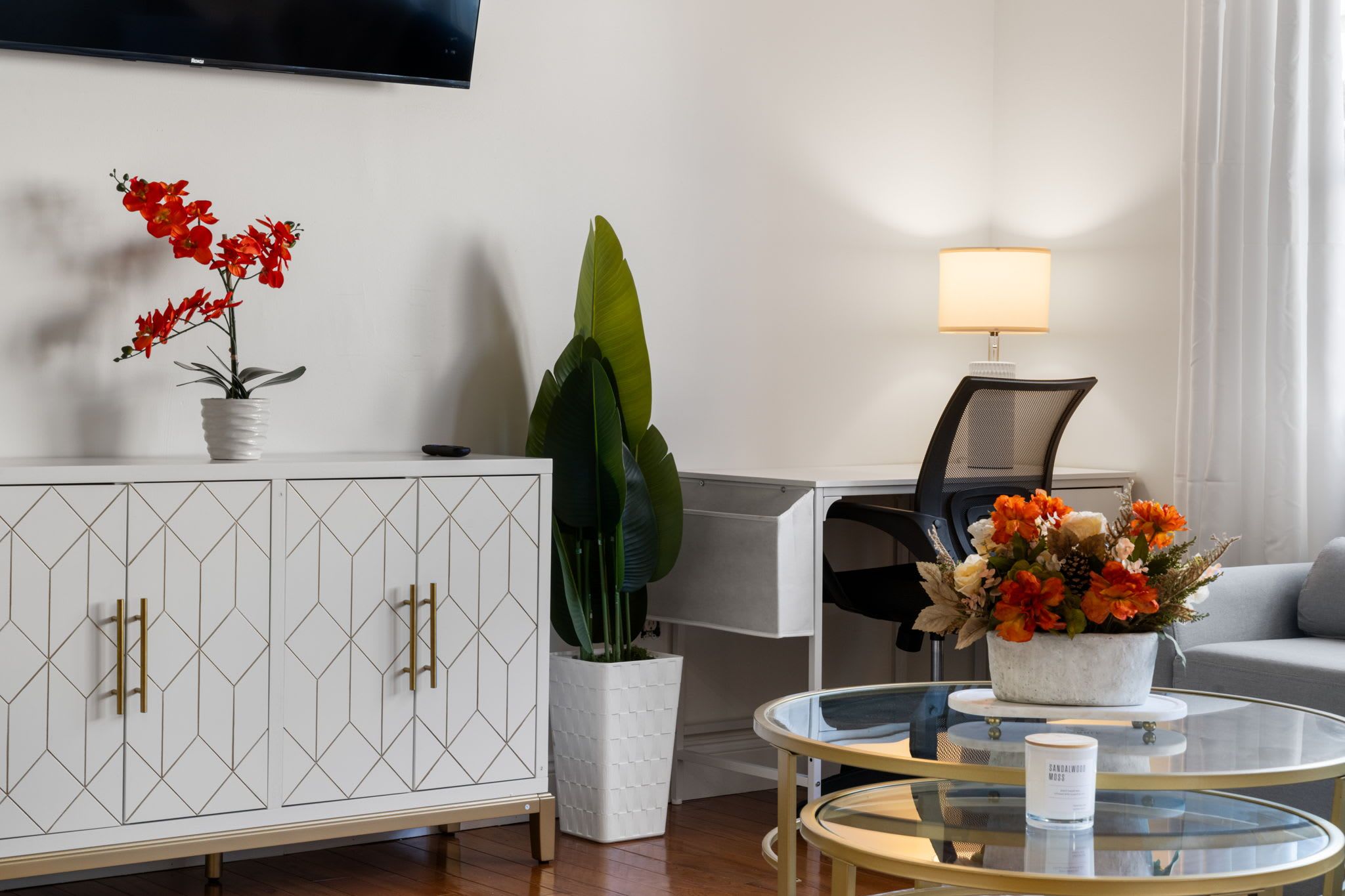 A modern living room with a geometric-patterned cabinet, a large green plant, a glass coffee table with a floral centerpiece, and a black office chair.