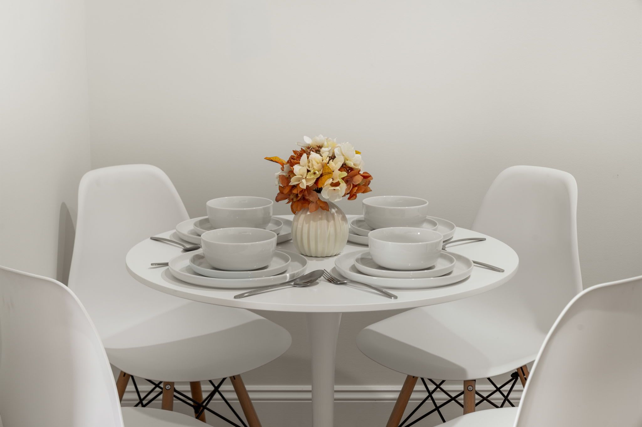 A white dining table with four chairs, a vase with flowers, and multiple white plates and bowls.