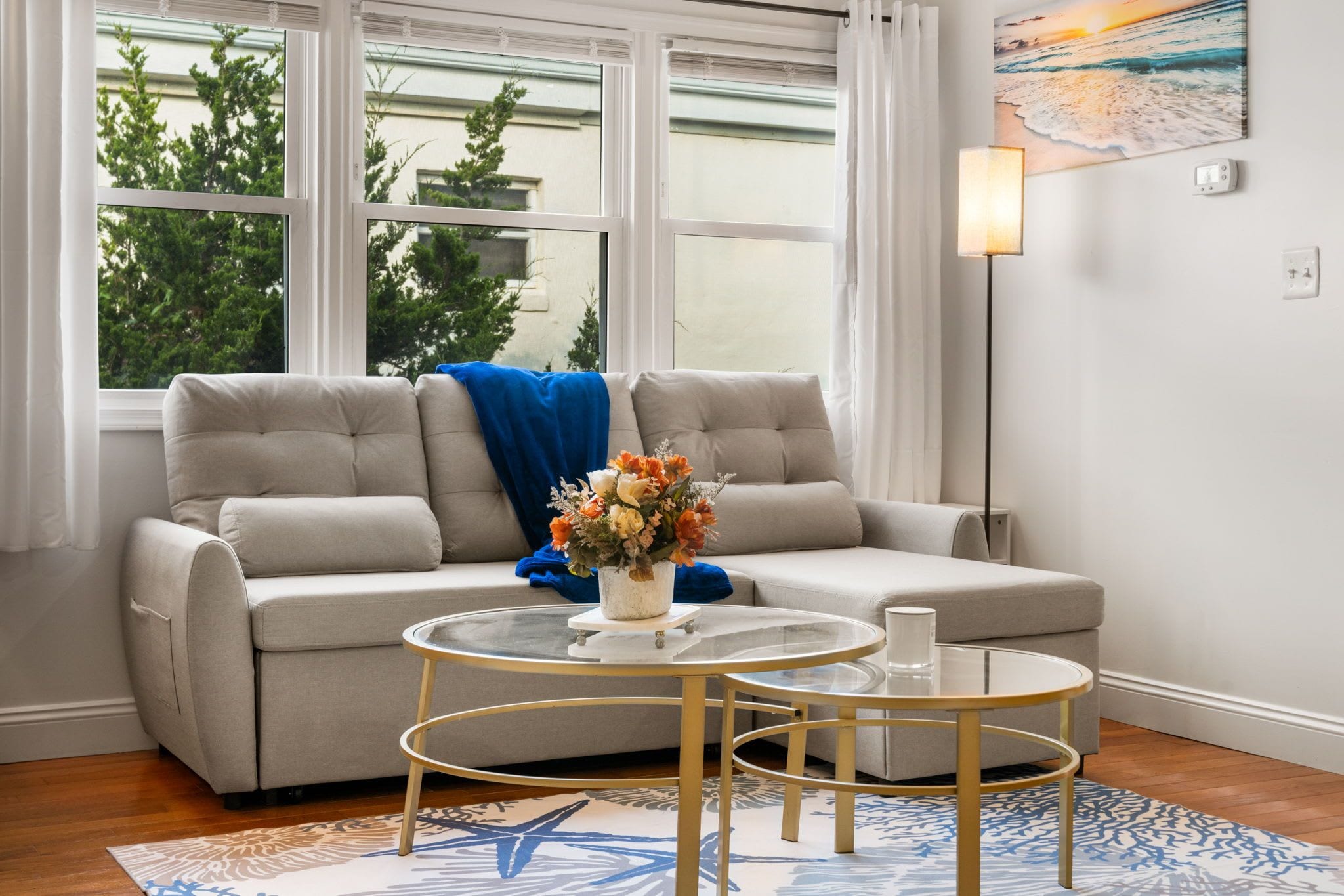 A living room with a sofa, two glass tables, a vase of flowers, and a painting on the wall.