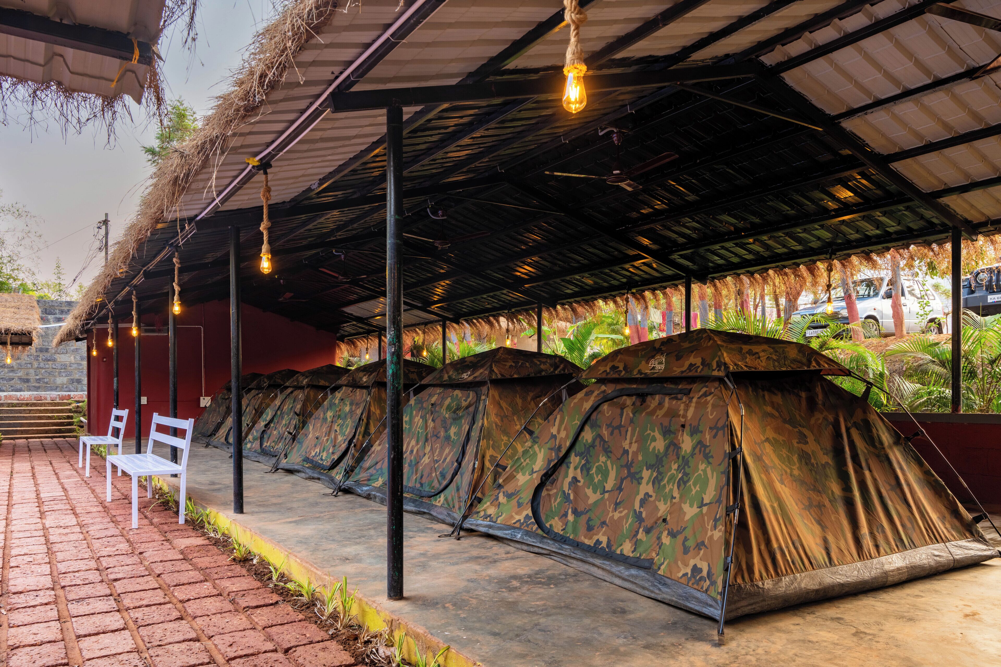 A row of camouflage tents under a covered area with hanging lights, a brick pathway, and a vehicle in the background.