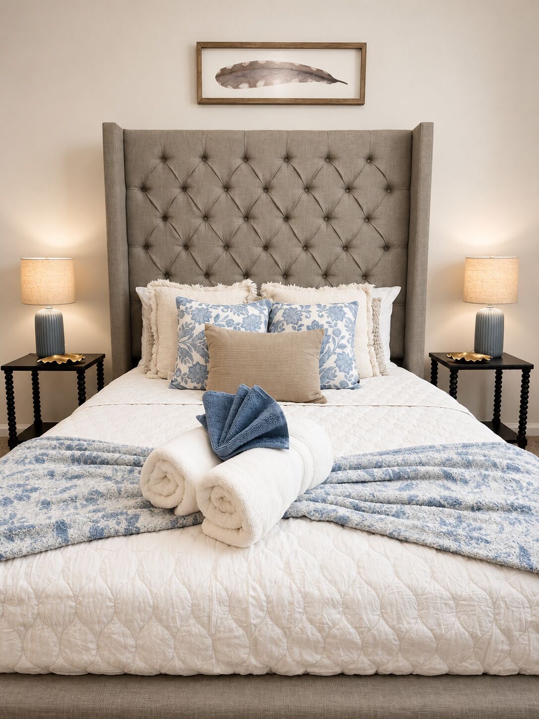 A neatly made bed with a tufted headboard, a decorative feather wall hanging, and two bedside lamps on matching nightstands.