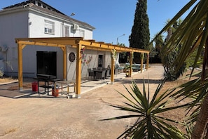 A covered outdoor seating area with tables and chairs, a building in the background, and a palm tree in the foreground. - Villa Perfecta: A 5-bedroom villa with a pool, just 30 minutes from Valencia! (Llíria)