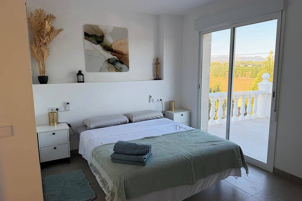 A bedroom with a bed, bedside tables, a nightstand, a window with a view of a balcony, and a wall-mounted artwork.