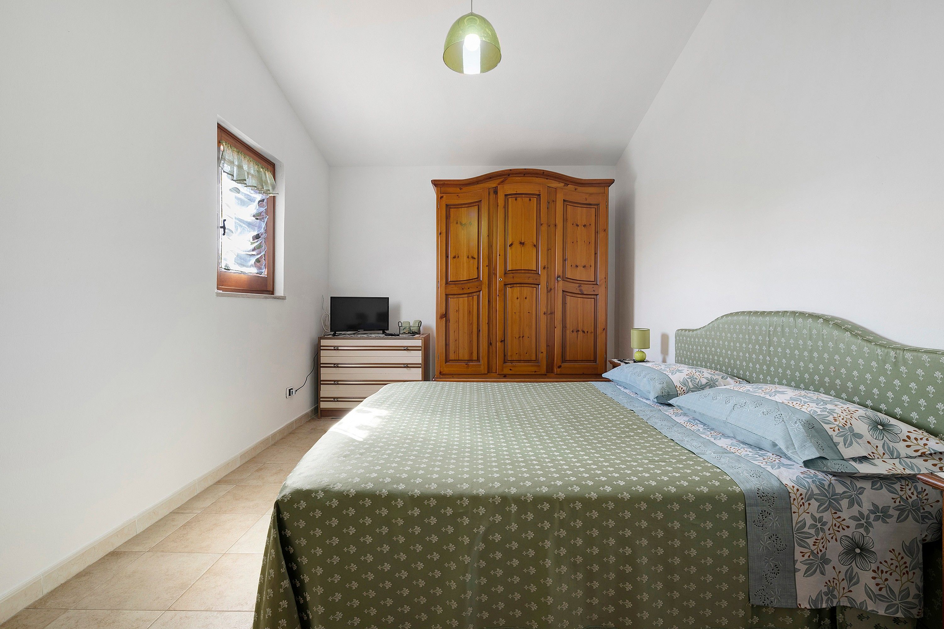 A bedroom with a bed, a wardrobe, a television on a dresser, and a window with curtains.