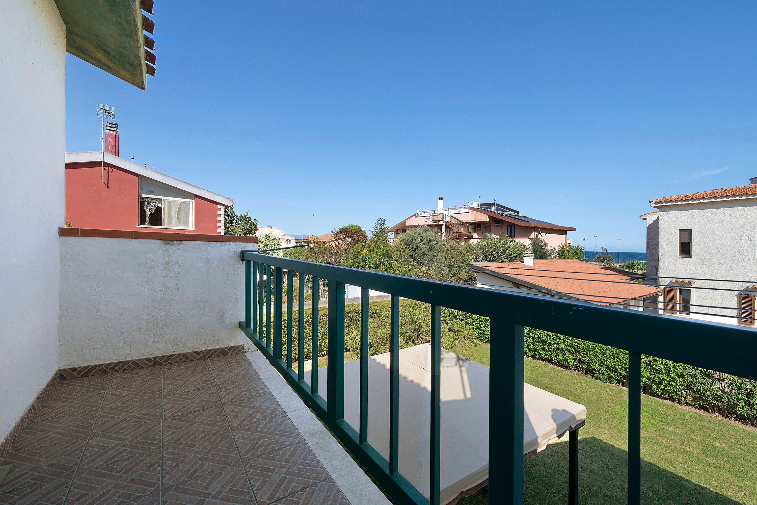Un balcone con ringhiera verde che si affaccia su una zona residenziale con case e cielo sereno.