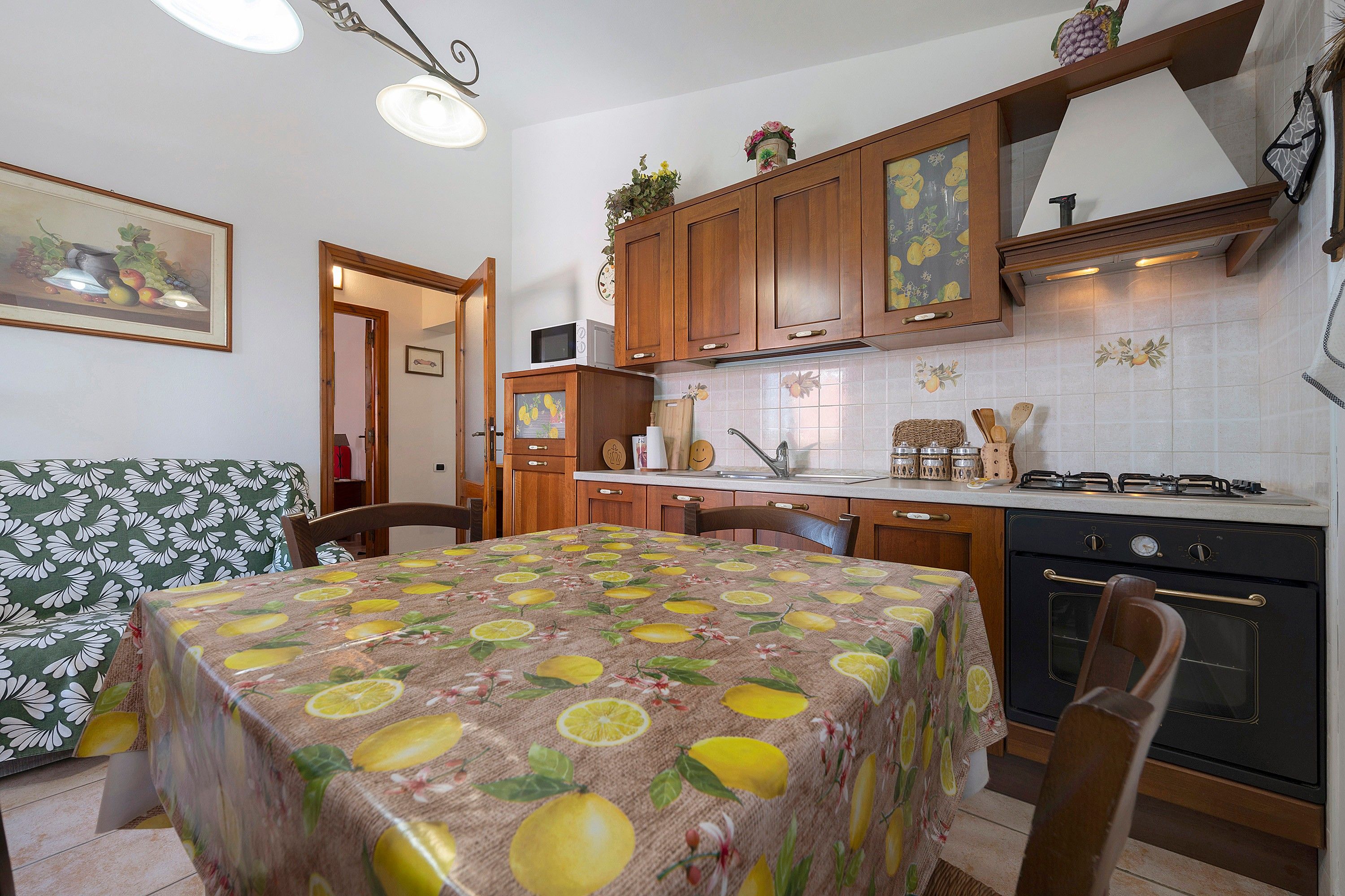 Een keuken met houten kastjes, een eettafel met een tafelkleed met een fruitprint, een fornuis en een magnetron.