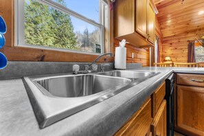 A modern kitchen with a double stainless steel sink, wooden cabinets, and a view of trees through a large window. - Cabin beside Shepherd Creek with Hot Tub and Fire Pit - A World’s Inn Property (Bryson City)