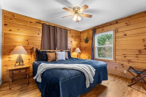 Ein Schlafzimmer mit einem Bett, zwei Nachttischlampen, einem Deckenventilator, einem Fenster mit Blick auf Bäume und Holzwänden.
