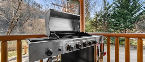 Una parrilla de gas portátil con varios controles, un tanque de propano y una barandilla de madera para terraza.