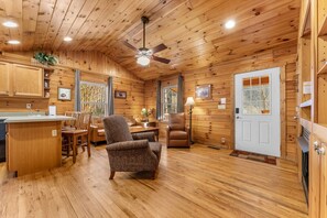 A cabin interior with a dining area, a living room with a sofa and armchair, a kitchen with wooden cabinets, and a ceiling fan. - Cabin beside Shepherd Creek with Hot Tub and Fire Pit - A World’s Inn Property (Bryson City)