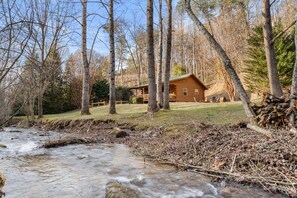 A cabin with a deck, surrounded by trees and a stream. - Cabin beside Shepherd Creek with Hot Tub and Fire Pit - A World’s Inn Property (Bryson City)