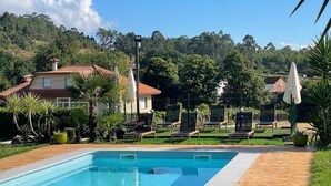 Piscina com espreguiçadeiras e guarda-sóis, cercada por vegetação e casas.
