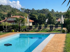 Ein Schwimmbecken mit Liegestühlen und Sonnenschirmen, umgeben von Grünflächen und Häusern.