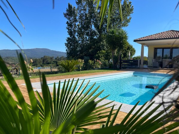 A swimming pool with a view of a mountain, surrounded by greenery and a building with a tiled roof.