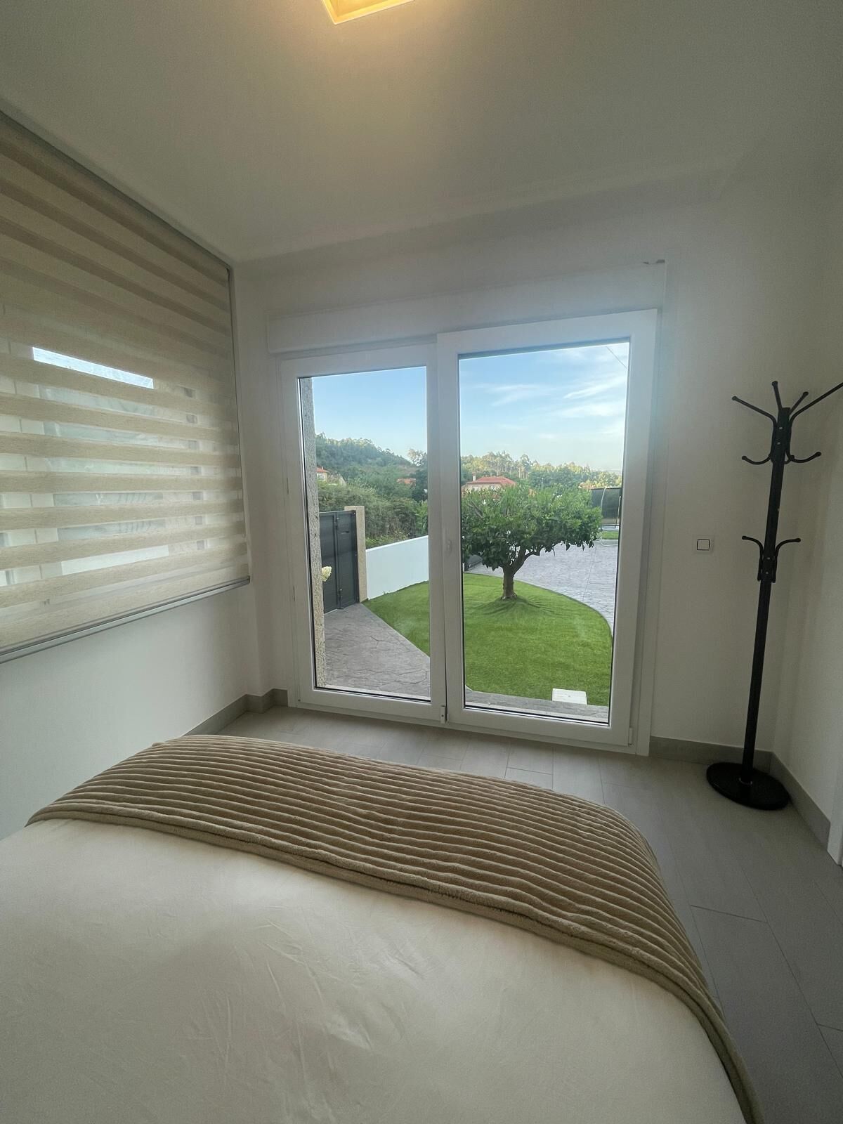 A bedroom with a bed, a coat rack, and a sliding glass door leading to a balcony with a view of a green lawn and trees.