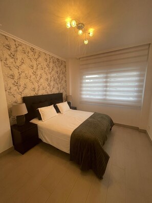 A bedroom with a bed, nightstands, a lamp, a ceiling light fixture, a window with blinds, and a patterned wall.
