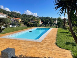 Piscina retangular com deck de madeira ao redor, com casas e vegetação ao fundo.