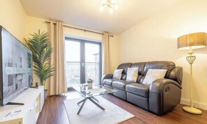 Ein Wohnzimmer mit einer Ledersofa, einem Glastisch, einem Fernseher auf einem Schrank, einer Stehlampe und einem großen Fenster mit Vorhängen.