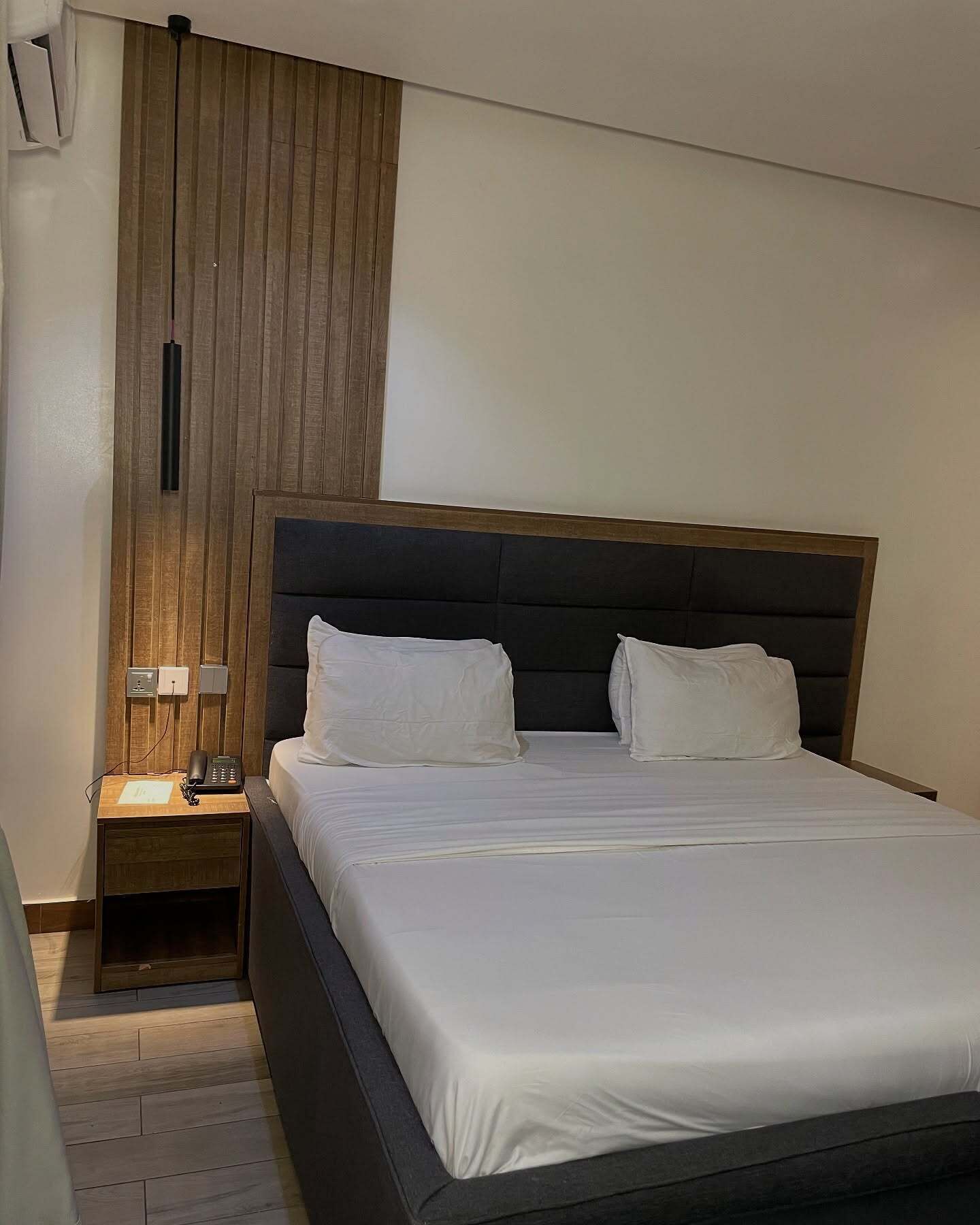 A neatly made bed with a dark headboard and white bedding, a bedside table with a lamp, and a wall-mounted air conditioning unit.