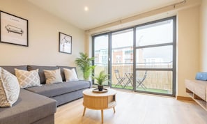 A modern living room with a grey sofa, a round wooden coffee table, and a large window offering a view of the outside. - Belfast Quarters: 5th Floor Gem! (Belfast)