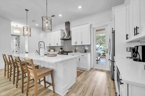 A modern kitchen with white cabinetry, a central island with bar stools, stainless steel appliances, and pendant lighting. - Lovely Day 30a by Stay on 30a (Inlet Beach)