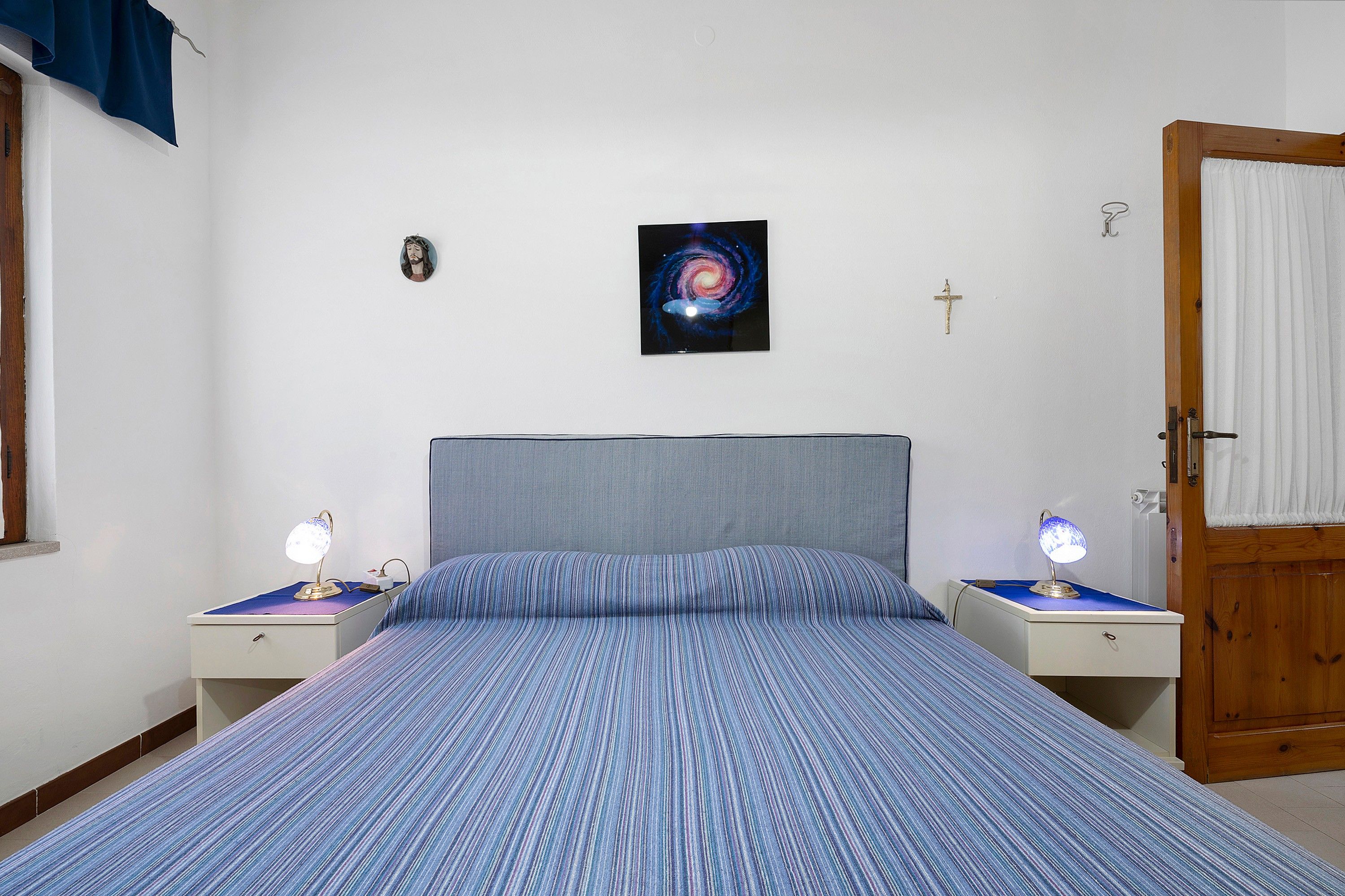 Une chambre à coucher avec un lit, deux tables de chevet avec des lampes, un tableau accroché au mur, une porte et une fenêtre avec des rideaux.