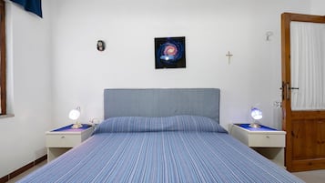 Une chambre à coucher avec un lit, deux tables de chevet avec des lampes, un tableau accroché au mur, une porte et une fenêtre avec des rideaux.